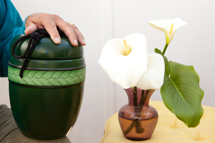 Urna funerária verde ao lado de um vaso com flores brancas, com uma pessoa apoiando a mão sobre a urna.