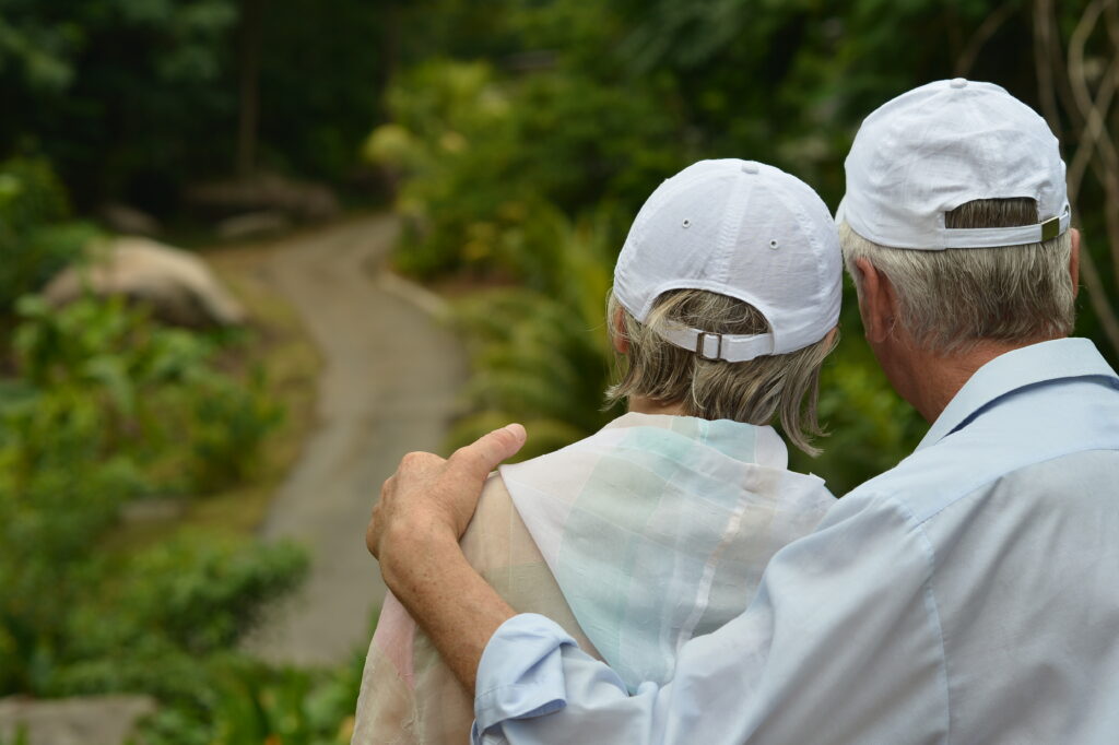 Casal de idosos de costas, abraçado em uma trilha, representando o companheirismo, o cuidado e a tranquilidade ao longo da vida.