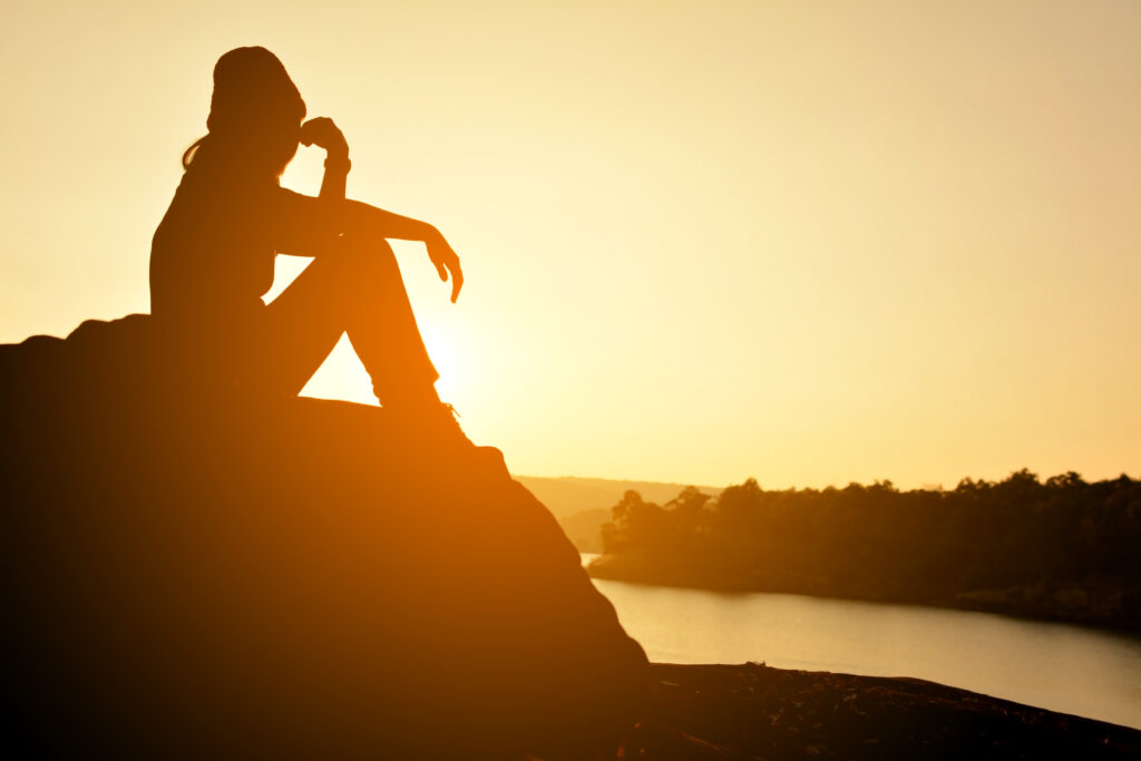 Silhueta de uma mulher sentada sobre uma rocha ao pôr do sol, contemplando a paisagem.